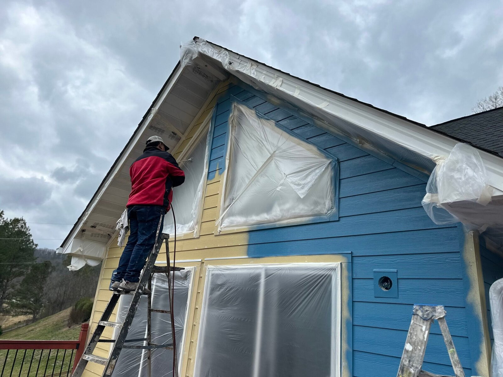 Painter on a ladder cutting in a gable, with the windows and the gutter masked off in plastic and the old yellow still showing beside the new blue