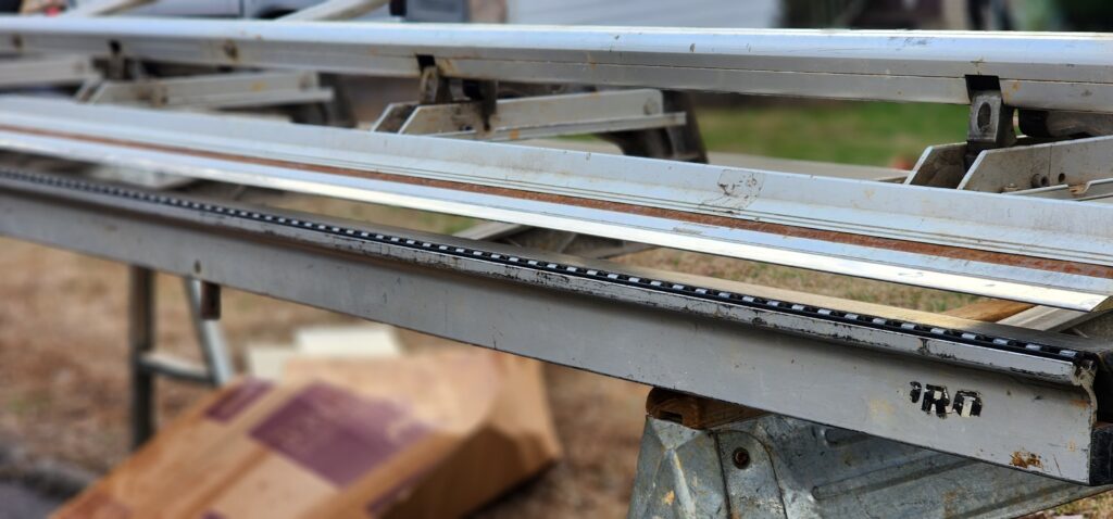 "A high-resolution photorealistic shot of a professional aluminum brake (bending machine) on a job site in Knoxville, Tennessee. A piece of white aluminum coil is being precisely bent to create a custom window wrap. In the background, a house with new siding is visible. The scene looks professional, organized, and technical. Natural daylight, 8k resolution, sharp focus on the metal textures."