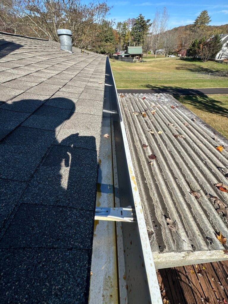 Open gutter along a shingle roof edge in Knoxville, TN with grime inside the run and fallen leaves on the porch roof below
