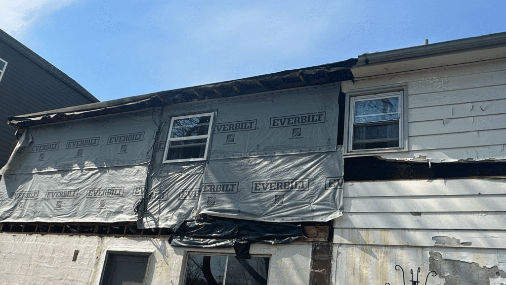 Siding torn away below the eave exposing housewrap and loose hanging boards