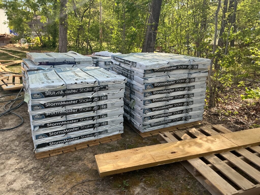 Stacked bundles of laminated shake-profile shingles on pallets in a wooded yard, delivered ahead of a roof replacement