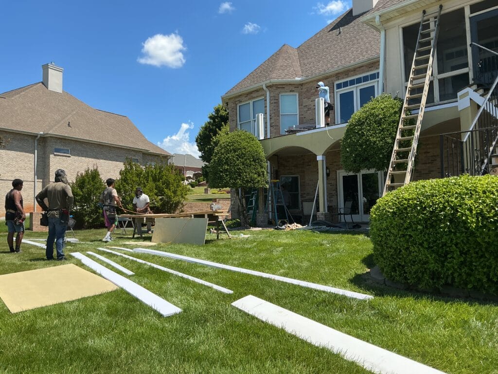 MI crew cutting trim on a work table outside a brick home, with ladders up to the second floor