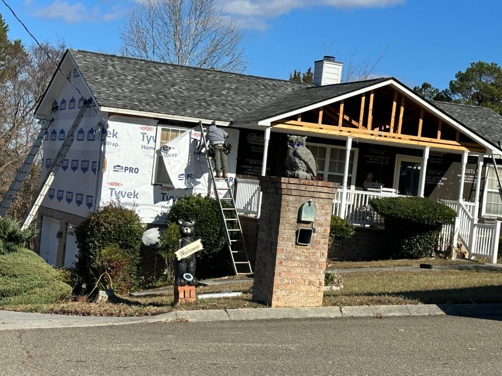 Ranch home part-way through a siding replacement: weather barrier exposed across the gable and upper wall while an installer works from a ladder, with new porch framing in place