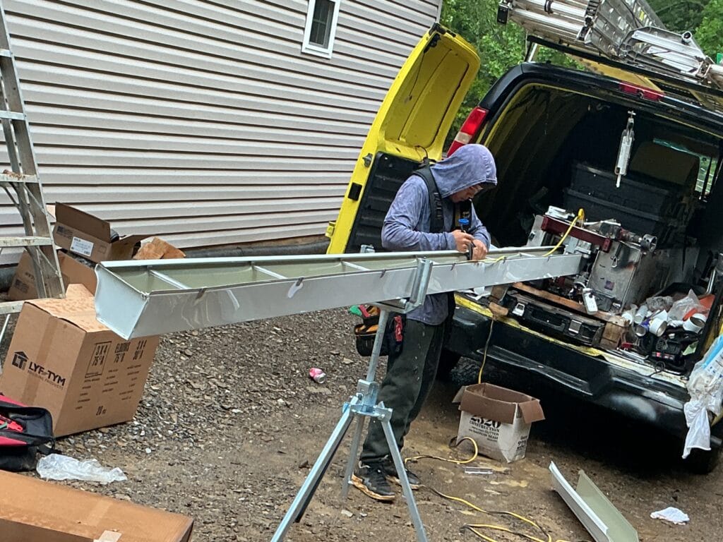 Installer forming a seamless gutter run on a roller stand at the back of the MI work van, with the forming machine inside and coil boxes on the ground