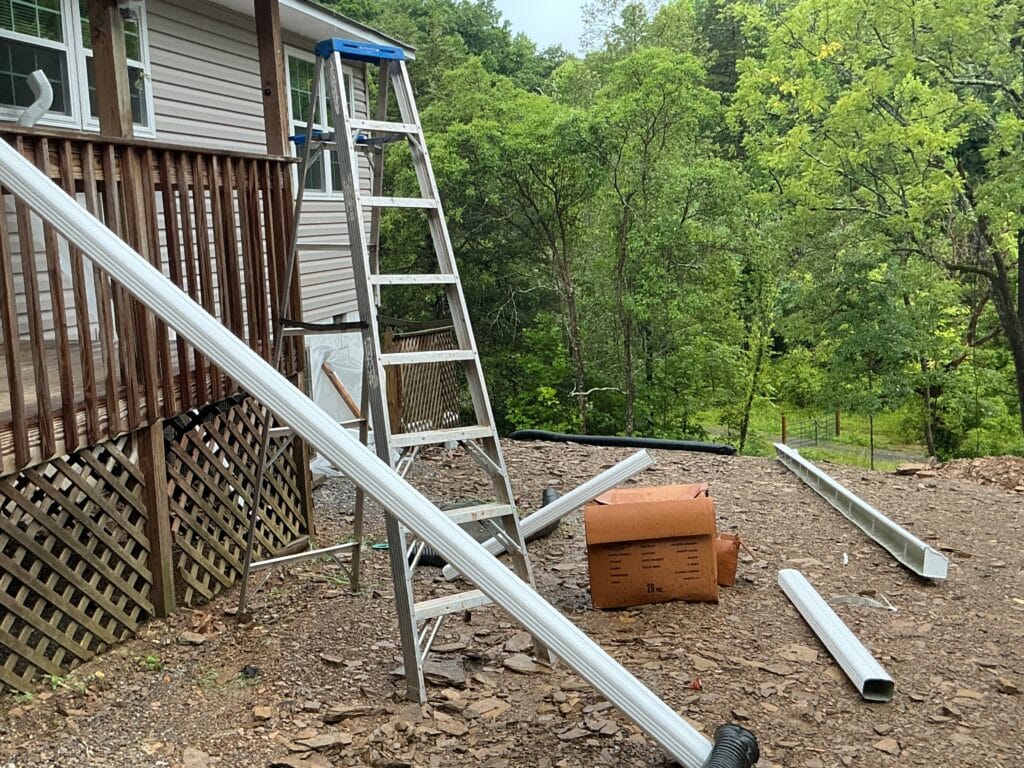 Formed gutter lengths and downspout stock laid out on a rocky hillside beside a step ladder, with dense woods rising behind the house