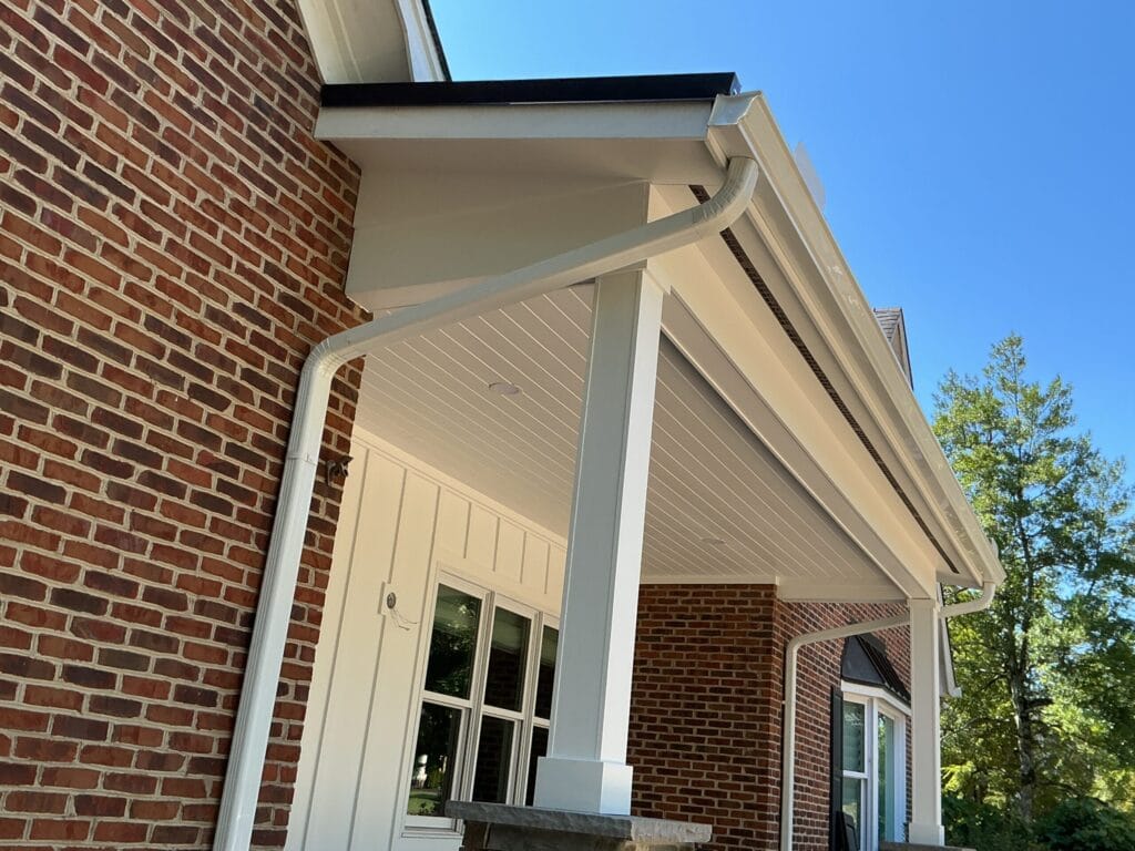 Finished white gutter running along a brick home’s porch eave, with the downspout elbow turning off the outlet and down the wall