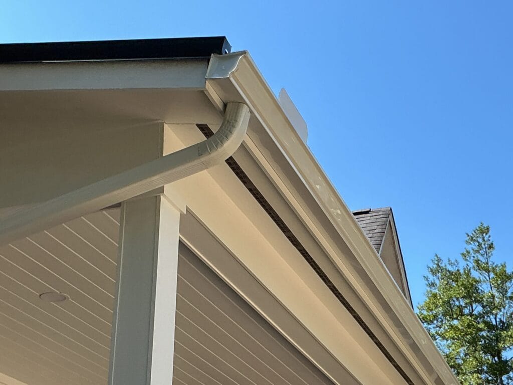 Roof eave from below: vented soffit panels, new fascia, and the gutter and downspout elbow that hang off it