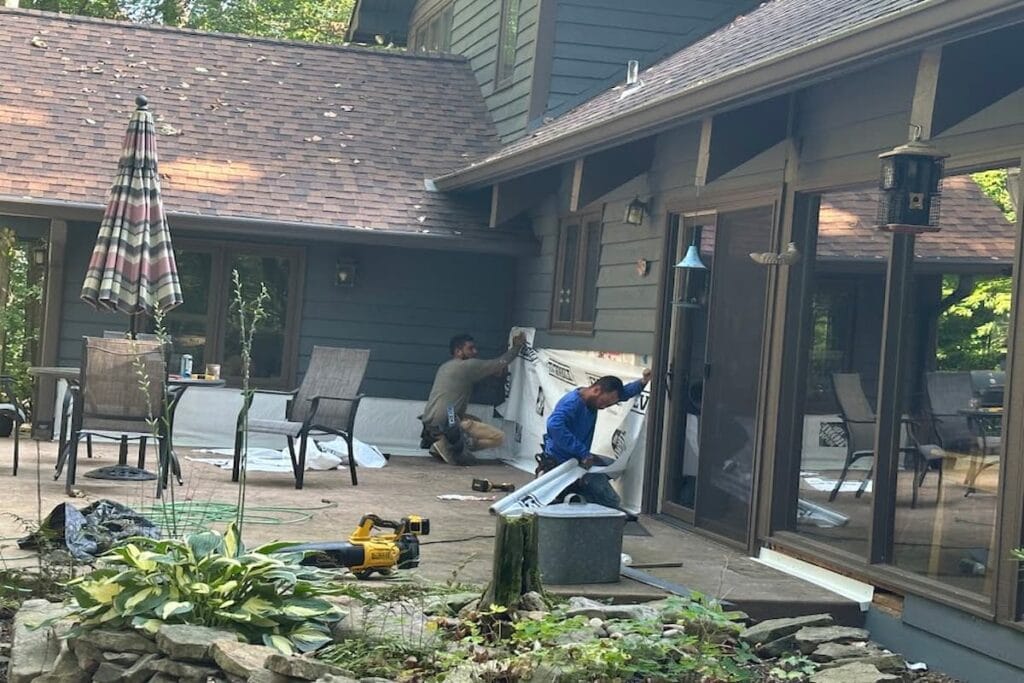 Two installers fitting weather barrier on the lower wall of a home on a wooded lot while the siding is repaired, with tools and drop sheets across the patio