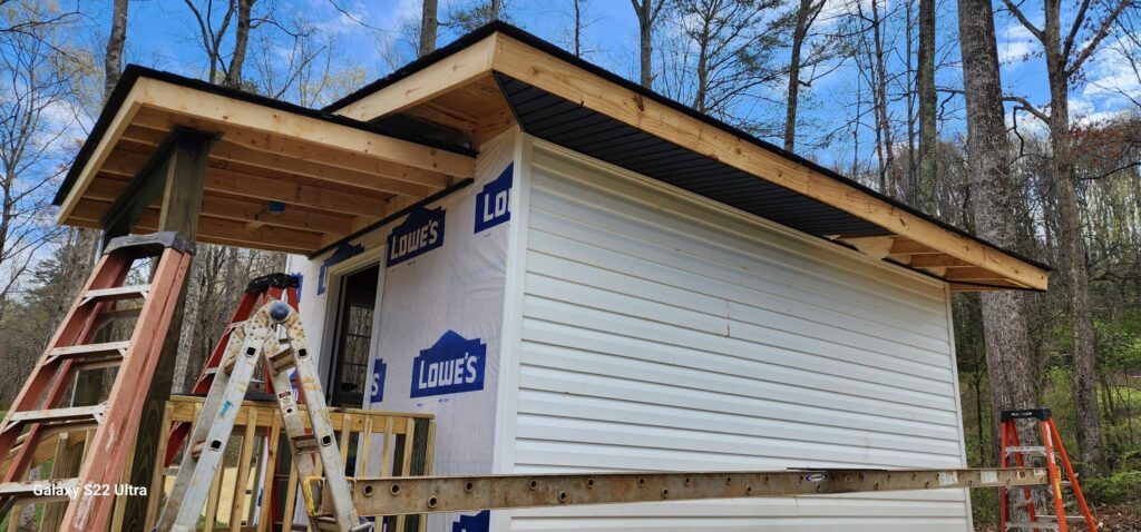 Small outbuilding under construction, with horizontal siding being installed over housewrap and a deep roof overhang