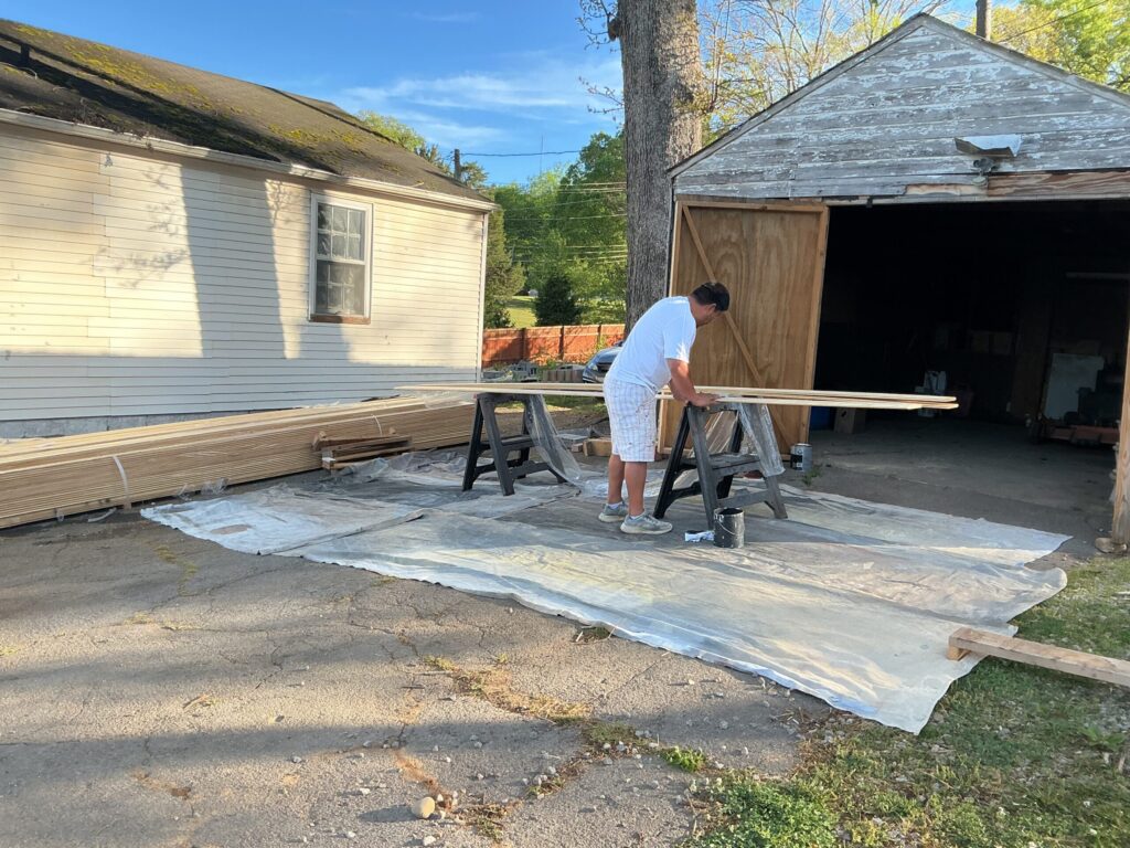 Worker handling long trim boards on sawhorses, with bundles of new boards on drop cloths and a weathered detached outbuilding behind